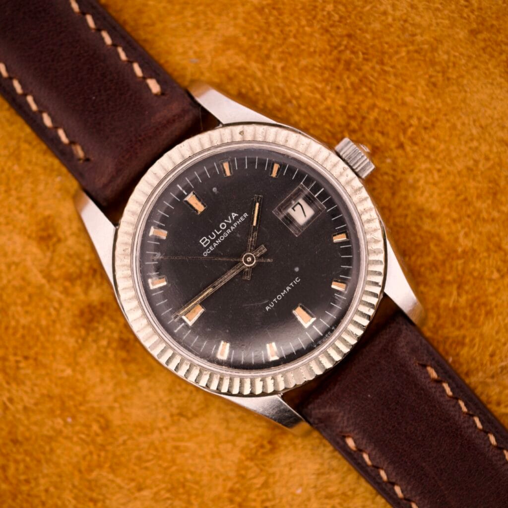 A Bulova Oceanographer Automatic Date with a black dial and fluted bezel sits atop a textured brown surface, paired with a brown leather strap.