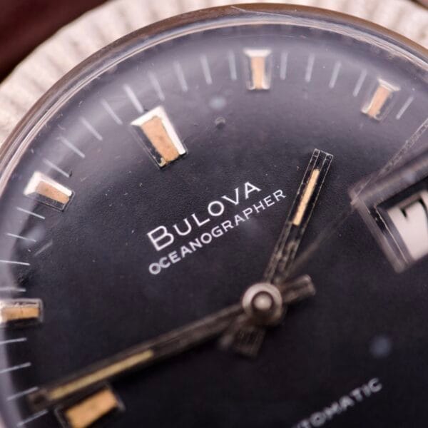 Close-up of the Bulova Oceanographer Automatic Date Black Dial Fluted Bezel watch face, highlighting the sleek black dial, visible hands, hour markers, and a portion of its automatic date window.