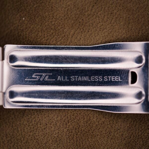 Close-up of a metal clasp engraved with STC ALL STAINLESS STEEL, set on brown textured background—an iconic detail from the Bulova Snorkel Automatic Devil Diver Ref. 386 Black Dial.