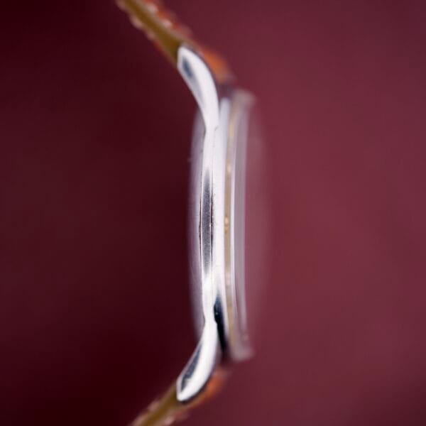 Side view of the Zenith Caltrava Jumbo with Arabic numerals, subseconds, a brown strap, and stainless steel case, set against a blurred maroon background.