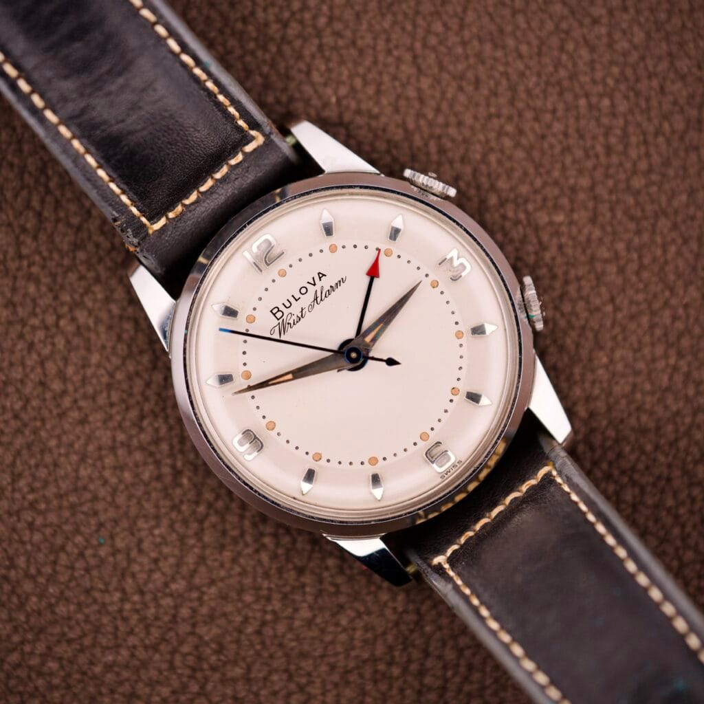 A NOS Bulova Wrist Alarm Manual Wind Cal. 11AERC with a white dial and brown leather strap rests on a textured brown surface.