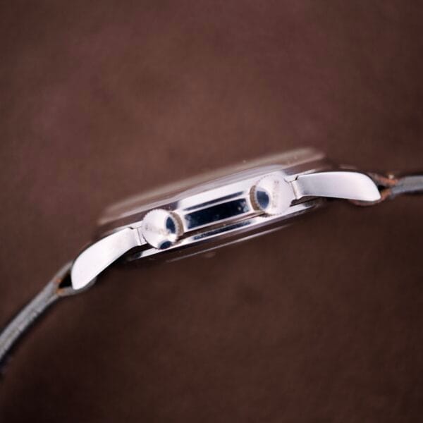 Side view of a NOS Bulova Wrist Alarm Manual Wind Cal. 11AERC with white dial, silver case, lugs, and leather strap on a brown background.