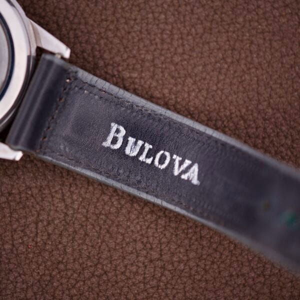 A close-up of a NOS Bulova Wrist Alarm Manual Wind Cal. 11AERC with a white dial, featuring its black strap on a brown textured surface.