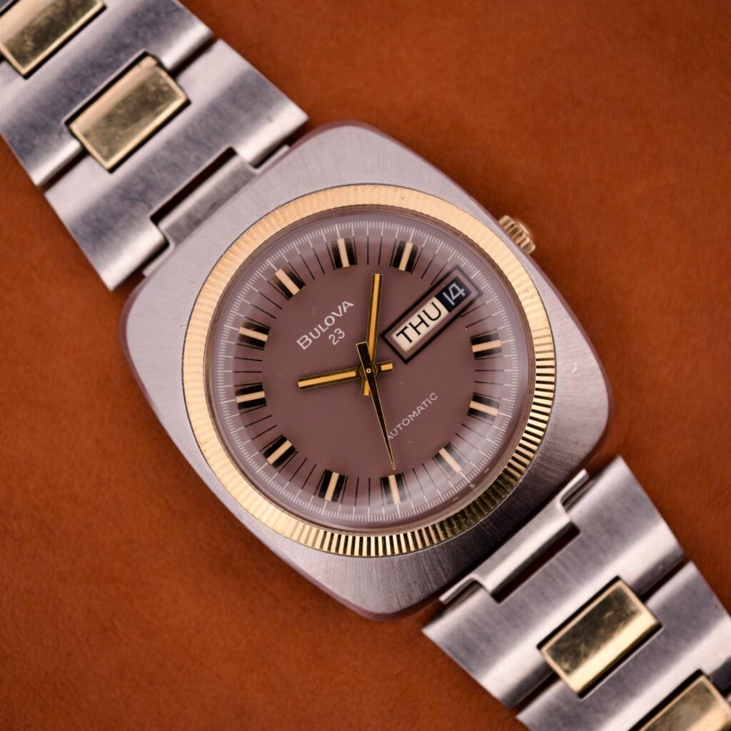A Bulova Day/Date 23 Jewels Automatic Two Tone Cushion Case Fluted Bezel wristwatch with a brown dial and day-date display rests on a brown surface.