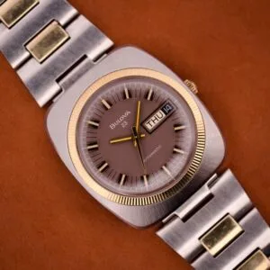 A Bulova Day/Date 23 Jewels Automatic Two Tone Cushion Case Fluted Bezel wristwatch with a brown dial and day-date display rests on a brown surface.