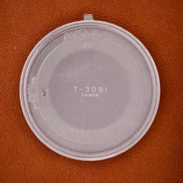 Round transparent plastic lid marked T-3081 Taiwan on a textured brown surface, possibly for a Bulova Day/Date 23 Jewels Automatic Two Tone Cushion Case Fluted Bezel watch.