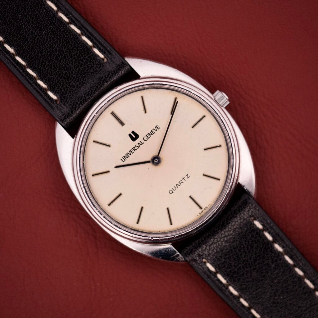 A Universal Genève Quartz Ref. 874 108 stainless steel cushion case watch with a cream dial and black leather strap rests on a red surface.