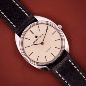 A Universal Genève Quartz Ref. 874 108 stainless steel cushion case watch with a cream dial and black leather strap rests on a red surface.