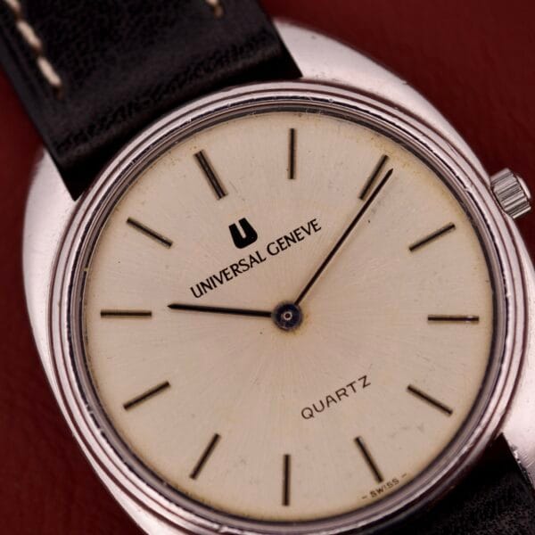 Close-up of a Universal Geneve Quartz Ref. 874 108 wristwatch featuring a stainless steel cushion case, cream dial, and black leather strap on a red surface.