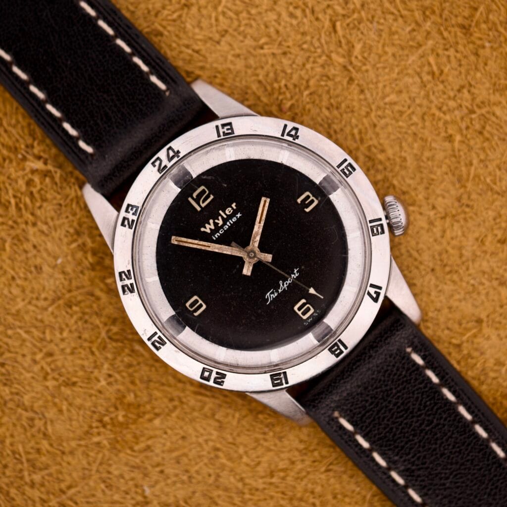 A Wyler Incaflex Tri Sport wristwatch with a stainless steel case and 24-hour bezel is displayed on a tan textured surface.