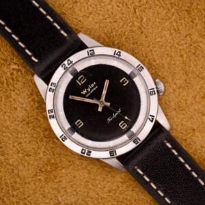 A Wyler Incaflex Tri Sport wristwatch with a stainless steel case and 24-hour bezel is displayed on a tan textured surface.