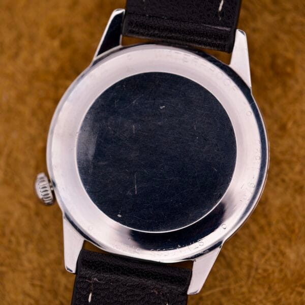 The back of a Wyler Incaflex Tri Sport Stainless Steel Case & 24 Hour Bezel wristwatch with a black strap is displayed on a brown textured surface.