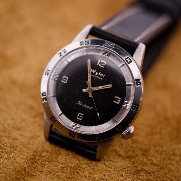 Close-up of a Wyler Incaflex Tri Sport wristwatch featuring a stainless steel case and 24-hour bezel, displayed on a brown surface.