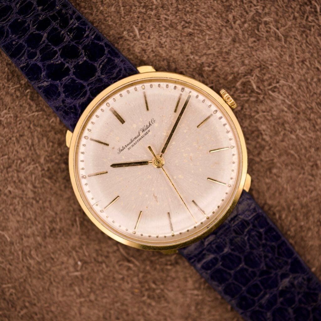 IWC "Stubby Lugs" Calatrava 18k Yellow Gold Cal. 401 with a white speckled dial and black leather strap, displayed on a brown textured surface.