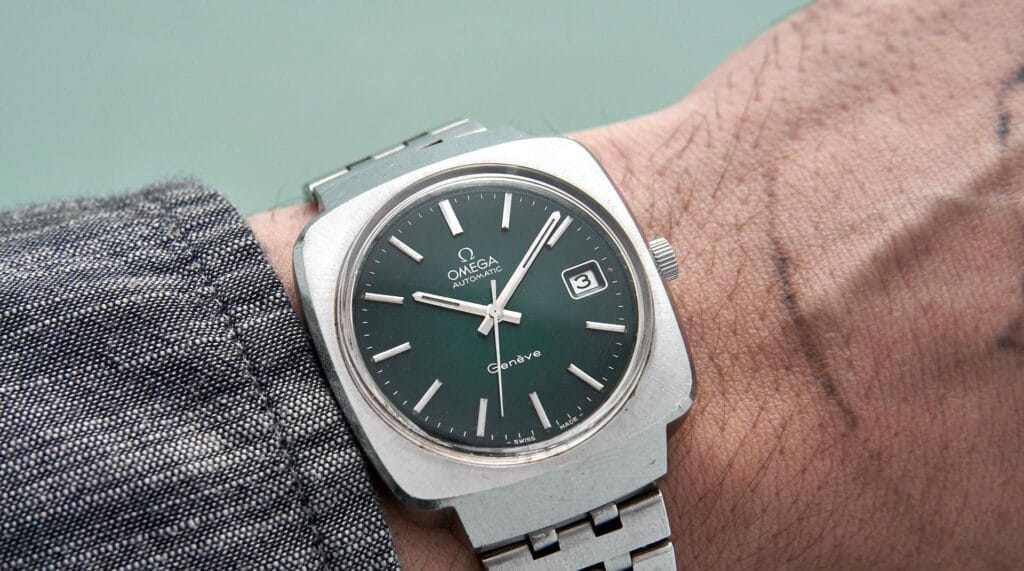 A silver Omega Genève wristwatch with a green dial is worn on a person’s left wrist.