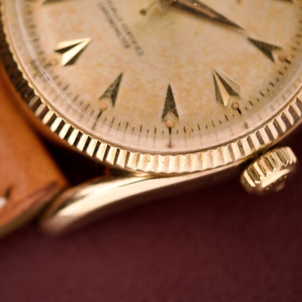 Close-up of a vintage Rolex Oyster Perpetual 'Bombay' Ref. 6593 with a 14K yellow gold case and tan leather strap, set against a rich burgundy background for an elegant touch.