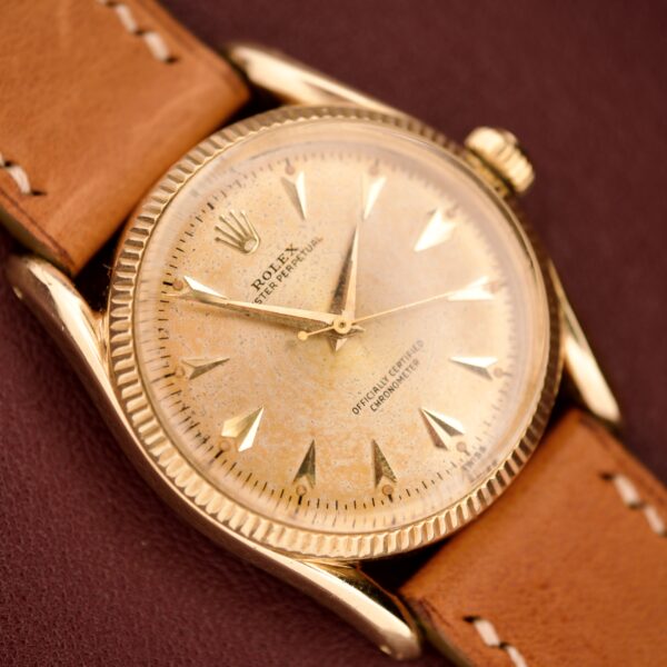 The Rolex Oyster Perpetual 'Bombay' Reference 6593, featuring a 14K yellow gold case and tan leather strap on a burgundy surface, captures the timeless elegance of Bombay's golden era.