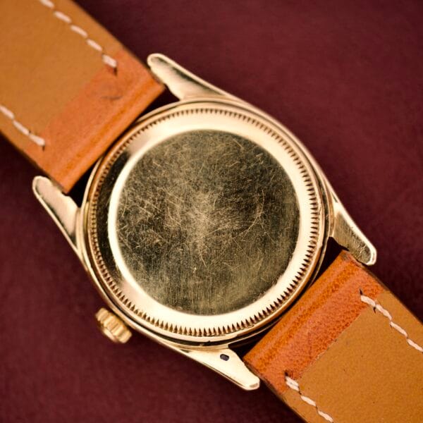A close-up of a scratched Rolex Oyster Perpetual 'Bombay' Reference 6593 14K yellow gold case back with tan leather straps on a maroon background, capturing a vintage vibe.