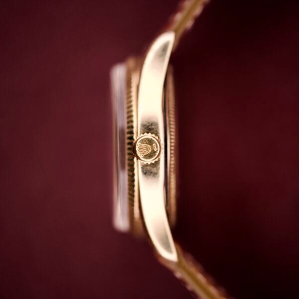 Close-up side view of the Rolex Oyster Perpetual 'Bombay' Ref. 6593's gold crown with logo, set against a blurred burgundy background—an elegant touch to this 14K yellow gold case timepiece.