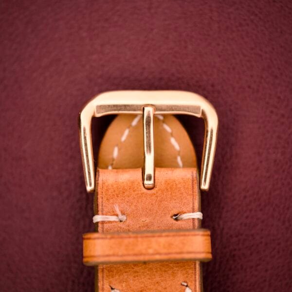 Close-up of a tan leather strap with gold buckle, echoing classic Rolex style, on a burgundy background. Shown with the Rolex Oyster Perpetual 'Bombay' Reference 6593 featuring a 14K yellow gold case.