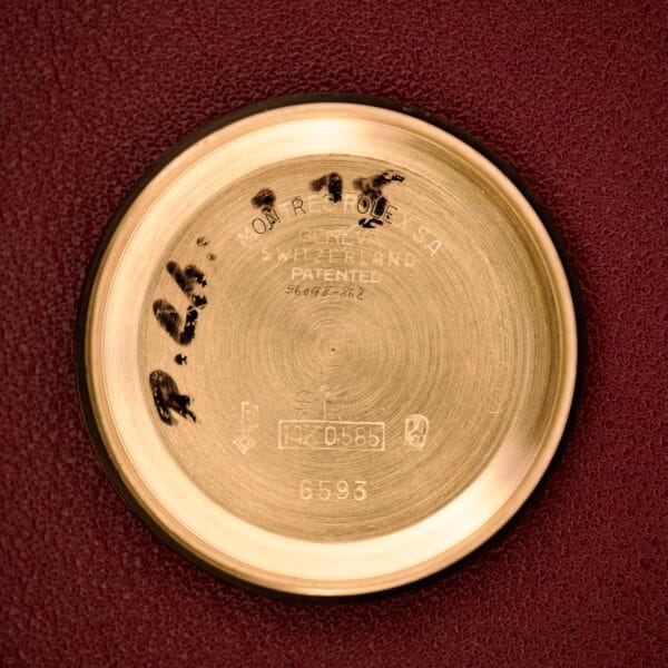 Rolex Oyster Perpetual 'Bombay' Ref. 6593 case back in 14K yellow gold shown on a red surface, engraved with Swiss patent details and displaying black smudge marks—classic Bombay design.