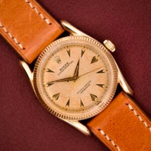 A Rolex Oyster Perpetual 'Bombay' Ref. 6593 with a 14K yellow gold case and tan leather strap is elegantly presented against a maroon backdrop, capturing the watch’s timeless Bombay sophistication.
