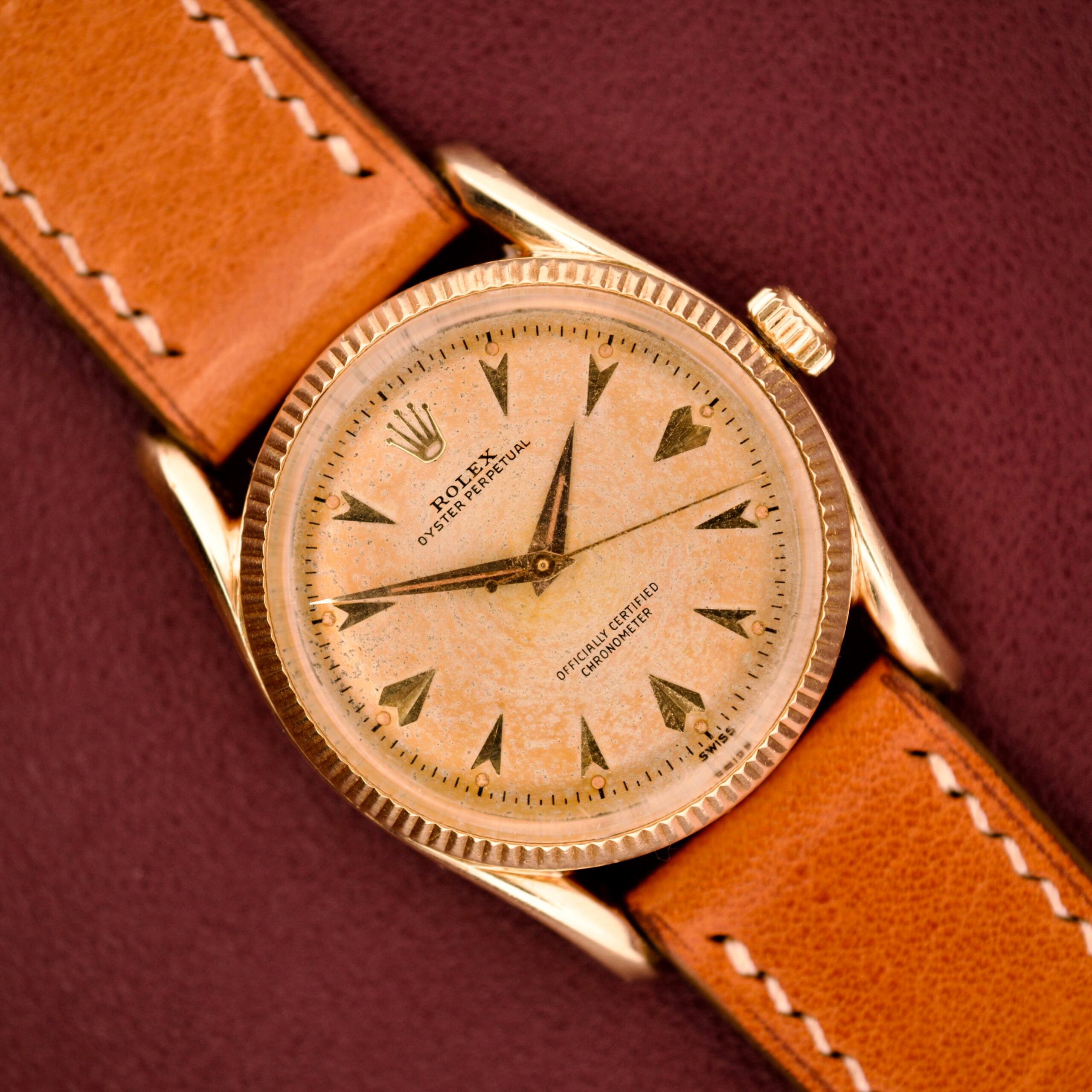 A Rolex Oyster Perpetual 'Bombay' Ref. 6593 with a 14K yellow gold case and tan leather strap is elegantly presented against a maroon backdrop, capturing the watch’s timeless Bombay sophistication. A Rolex Oyster Perpetual 'Bombay' Ref. 6593 with a 14K yellow gold case and tan leather strap is elegantly presented against a maroon backdrop, capturing the watch’s timeless Bombay sophistication.