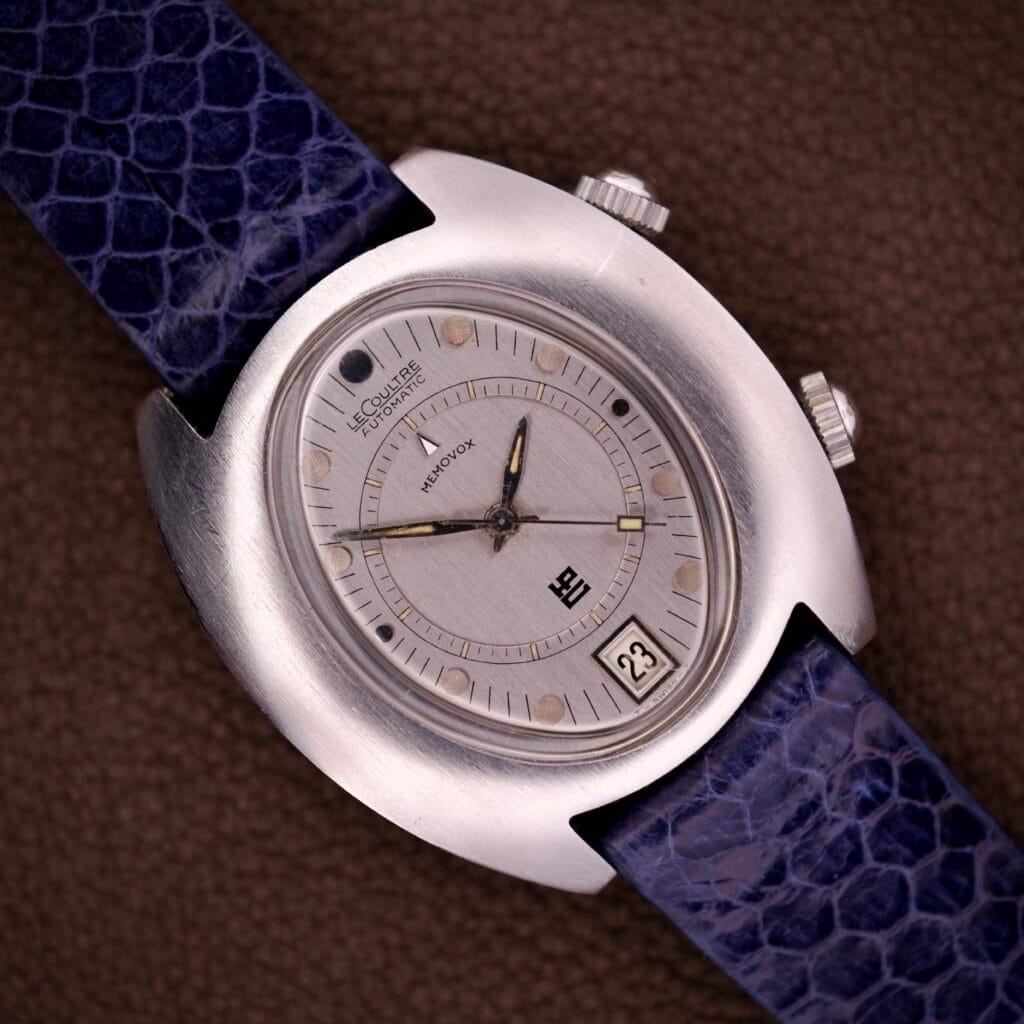 The Lecoultre Automatic Memovox Reveil "Egg" Ref. 3072-916 Cal. 916 wristwatch with a blue textured strap rests on a brown surface at 11:11, featuring the renowned Cal. 916 movement for exceptional precision and elegance.