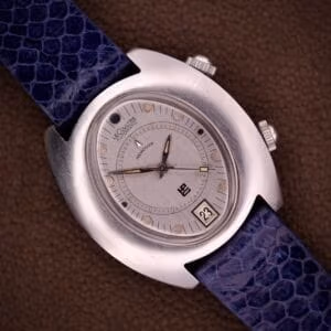 The Lecoultre Automatic Memovox Reveil "Egg" Ref. 3072-916 Cal. 916 wristwatch with a blue textured strap rests on a brown surface at 11:11, featuring the renowned Cal. 916 movement for exceptional precision and elegance.