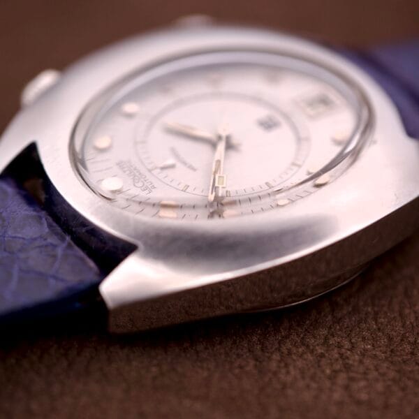 Lecoultre Automatic Memovox Reveil "Egg" Ref. 3072-916 Cal. 916 wristwatch with a silver case and blue textured strap, displayed close-up and angled on a brown surface.