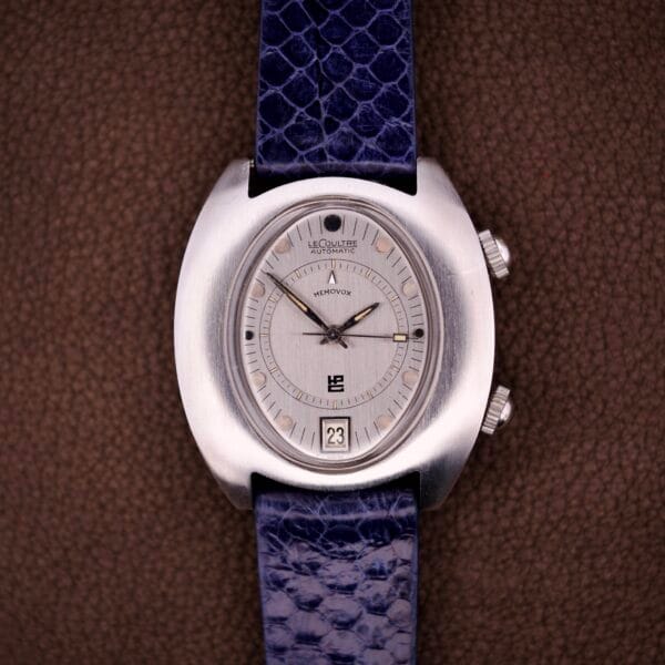 Silver vintage Lecoultre Automatic Memovox Reveil "Egg" Ref. 3072-916 wristwatch with blue textured strap, showcasing date and time; powered by the renowned Cal. 916 movement on a brown background.