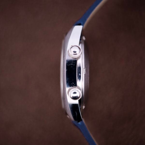 Side view of the Lecoultre Automatic Memovox Reveil "Egg" Ref. 3072-916 Cal. 916 wristwatch with blue strap and two round buttons, shown against a brown background.