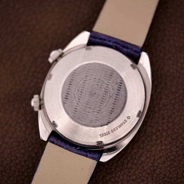 The back of a Lecoultre Automatic Memevox Reveil "Egg" Ref. 3072-916 Cal. 916 stainless steel watch with leather straps rests on a brown textured surface.