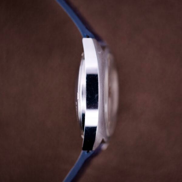 Side view of a Lecoultre Automatic Memevox Reveil "Egg" Ref. 3072-916 Cal. 916 wristwatch with a blue strap and silver case, shown against a brown background.