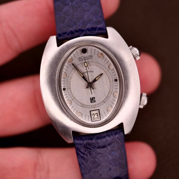 A Lecoultre Automatic Memovox Reveil "Egg" Ref. 3072-916 Cal. 916 silver wristwatch with a blue strap is held between fingers, showing the time and date.