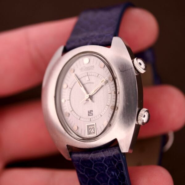 A hand holds a silver Lecoultre Automatic Memovox Reveil "Egg" Ref. 3072-916 Cal. 916 wristwatch with a blue strap and two crowns on the right.