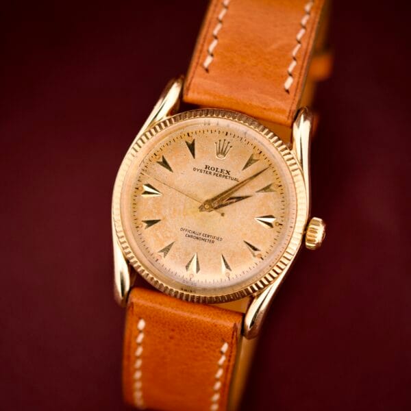 Rolex Oyster Perpetual 'Bombay' Ref. 6593 with a 14K yellow gold case and tan leather strap, elegantly showcased against a dark maroon background—an iconic timepiece inspired by Bombay’s timeless charm.