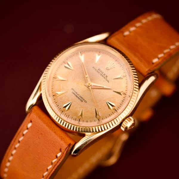 A Rolex Oyster Perpetual 'Bombay' Ref. 6593 with a 14K yellow gold case and tan leather strap is displayed on a dark red background, radiating timeless elegance and Bombay-inspired sophistication.