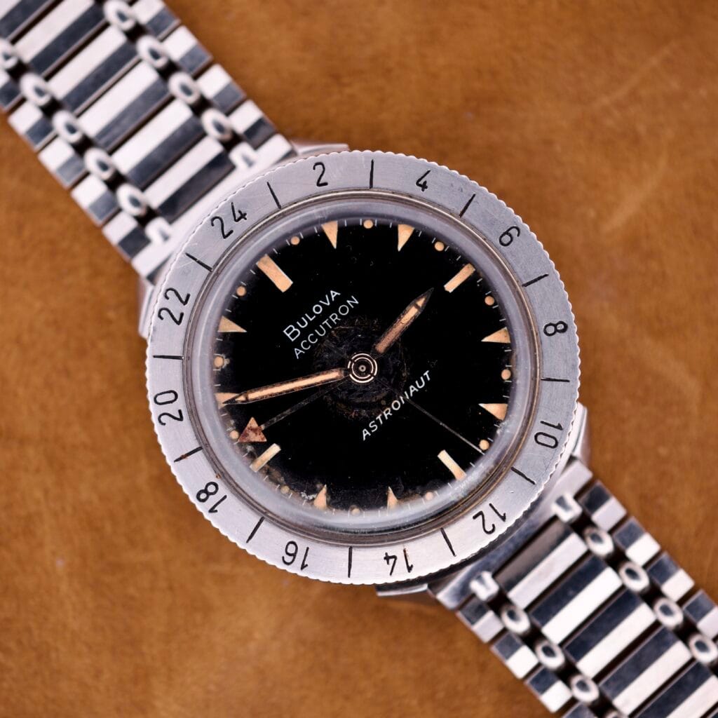 Bulova Accutron Astronaut 214 GMT 24 Hour Bezel Black Dial wristwatch with metal band, displayed on a brown surface.