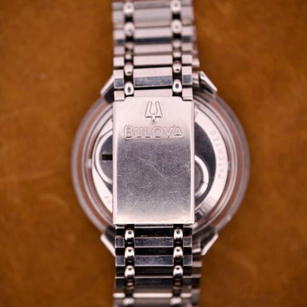 Close-up of a silver Bulova Accutron Astronaut 214 GMT 24 Hour Bezel Black Dial watch clasp and bracelet on a brown surface.