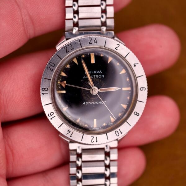 A hand holds a Bulova Accutron Astronaut 214 GMT 24 Hour Bezel wristwatch with a black dial and metal band.