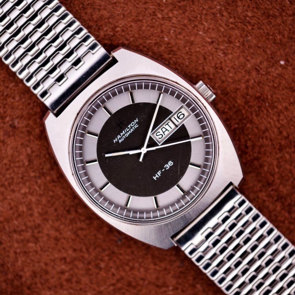 A Hamilton Automatic HF-36 Day-Date Hi-Beat Cal. 631 wristwatch with a metal band, displaying the day and date, rests on a brown textured surface.