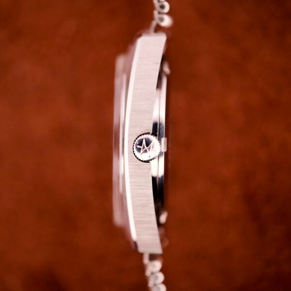 Side view of a Hamilton Automatic HF-36 Day-Date Hi-Beat Cal. 631 silver wristwatch with crown featuring a star logo, set against a blurred brown background.