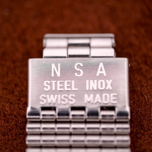 Close-up of a metal clasp engraved with NSA STEEL INOX SWISS MADE on a brown textured background—an ideal accessory for your Hamilton Automatic HF-36 Day-Date Hi-Beat Cal. 631.
