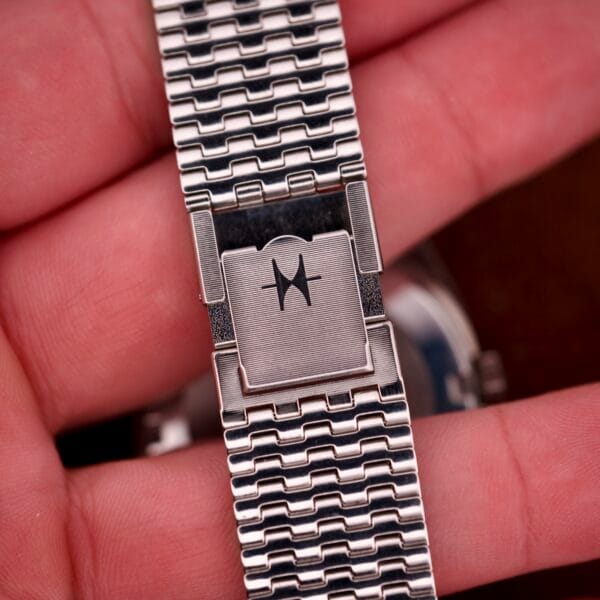 A close-up of a hand holding a metal watch band with a clasp engraved with an abstract logo, evoking the precision of the Hamilton Automatic HF-36 Day-Date Hi-Beat Cal. 631.