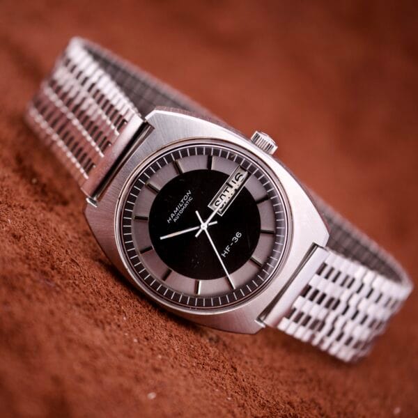 A Hamilton Automatic HF-36 Day-Date Hi-Beat Cal. 631 wristwatch with a silver case, black dial, and metal band featuring a Day-Date display rests on a brown textured surface.