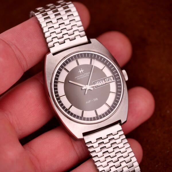 A hand holds a Hamilton Automatic HF-36 Day-Date Hi-Beat Cal. 631 wristwatch with a silver metal band and day-date display.