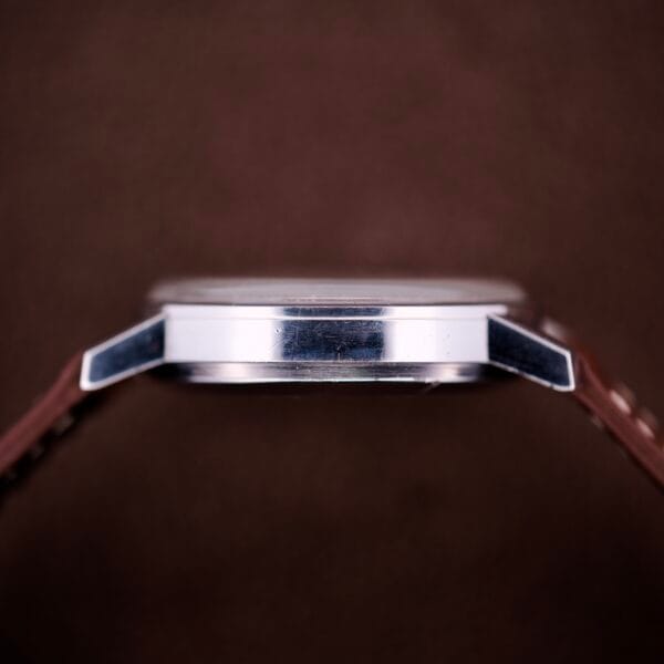 Side view of the Omega Automatic Seamaster Ref. 166.020 For "U.S. Steel" Cal. 1010 Full Set, with a silver case and brown strap, shown against a plain brown background.