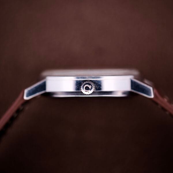 Side view of an Omega Automatic Seamaster Ref. 166.020 for "U.S. Steel" Cal. 1010 full set wristwatch with a brown strap and silver crown, displayed against a brown background.