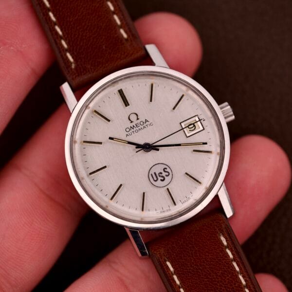 A hand holds an Omega Automatic Seamaster Ref. 166.020 for "U.S. Steel," featuring a brown leather strap, white dial, date display, and Cal. 1010 movement—full set included.